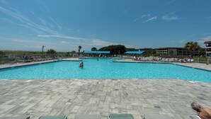 Ein Pool mit Liegestühlen und Sonnenschirmen, umgeben von Gebäuden und wolkenlosem Himmel.