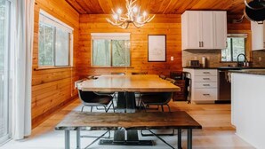 A wooden cabin interior with a dining table, chairs, a chandelier, and a kitchen area. - Zen Hideaway - Fiber - EV - King bed - Pet - Baker (Deming)