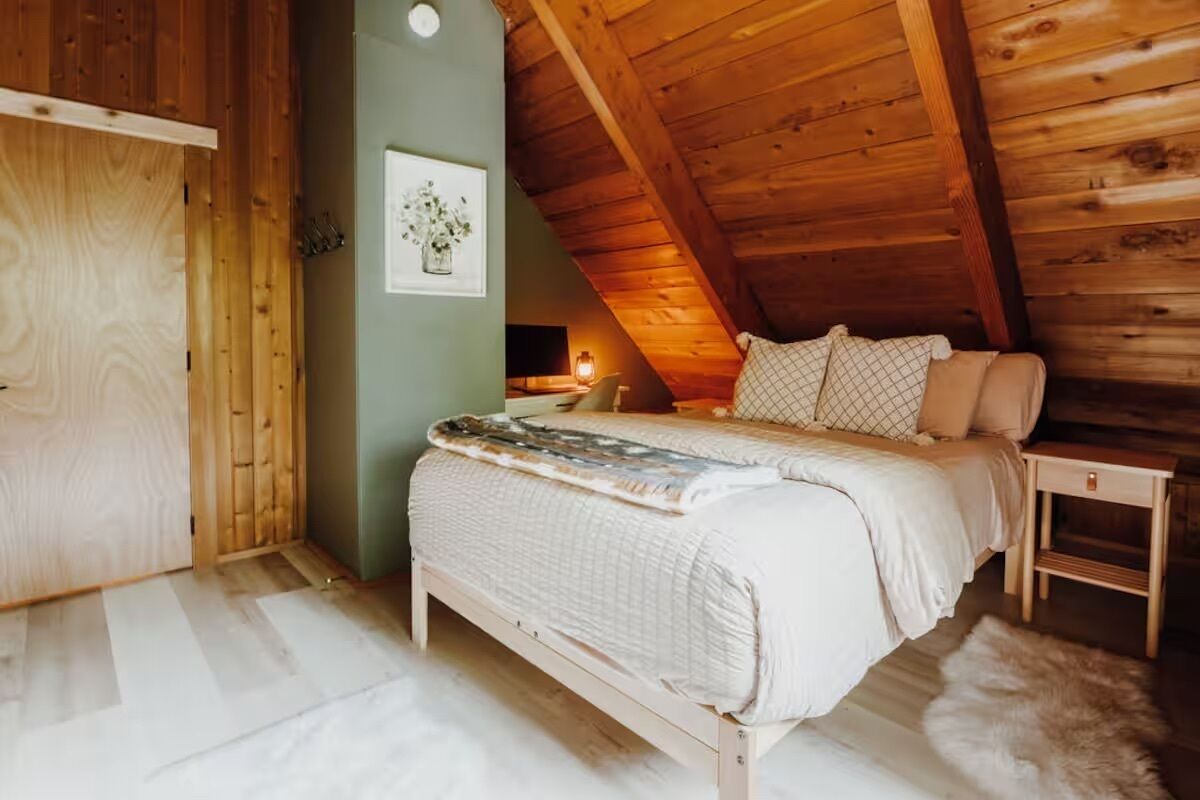 A cozy attic bedroom with a wooden ceiling, a bed with a quilted blanket, a nightstand, and a wall-mounted picture.