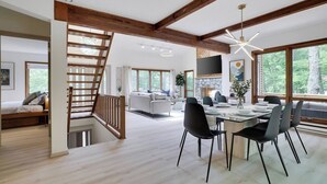 Amplia sala de estar con una escalera de madera, una mesa de comedor con sillas y grandes ventanales que ofrecen vistas al exterior.