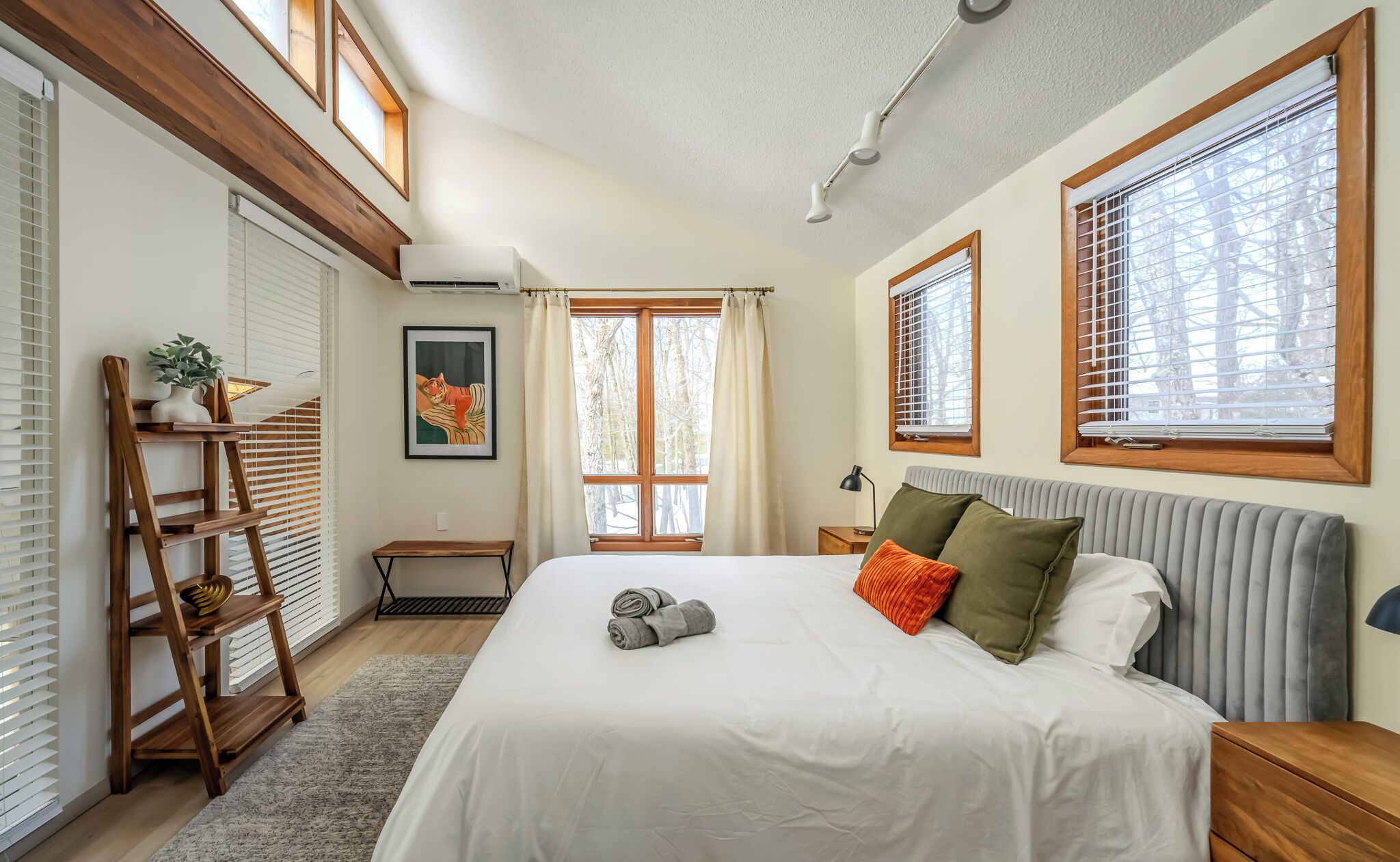A bedroom with a large bed, a wooden ladder, a small table, and a window with blinds.