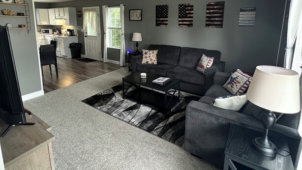 Ein Wohnzimmer mit Sofa, Couchtisch und Lampe, im Hintergrund eine Küche und ein Deckenventilator.