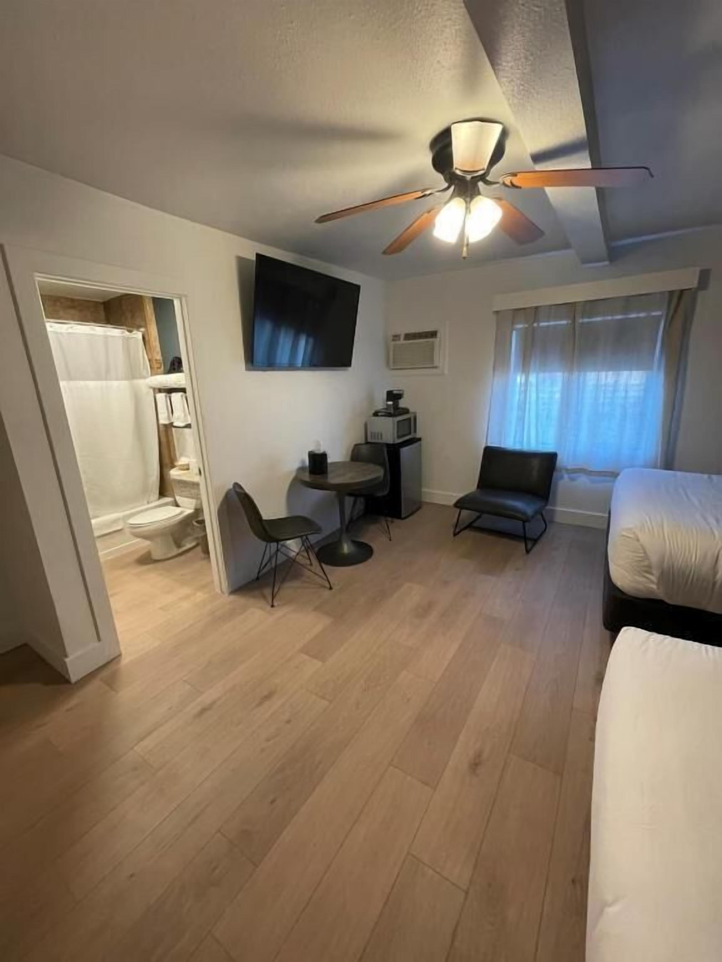 A compact hotel room with a ceiling fan, a small desk with a chair, a bed, a shower area, and a window with curtains.