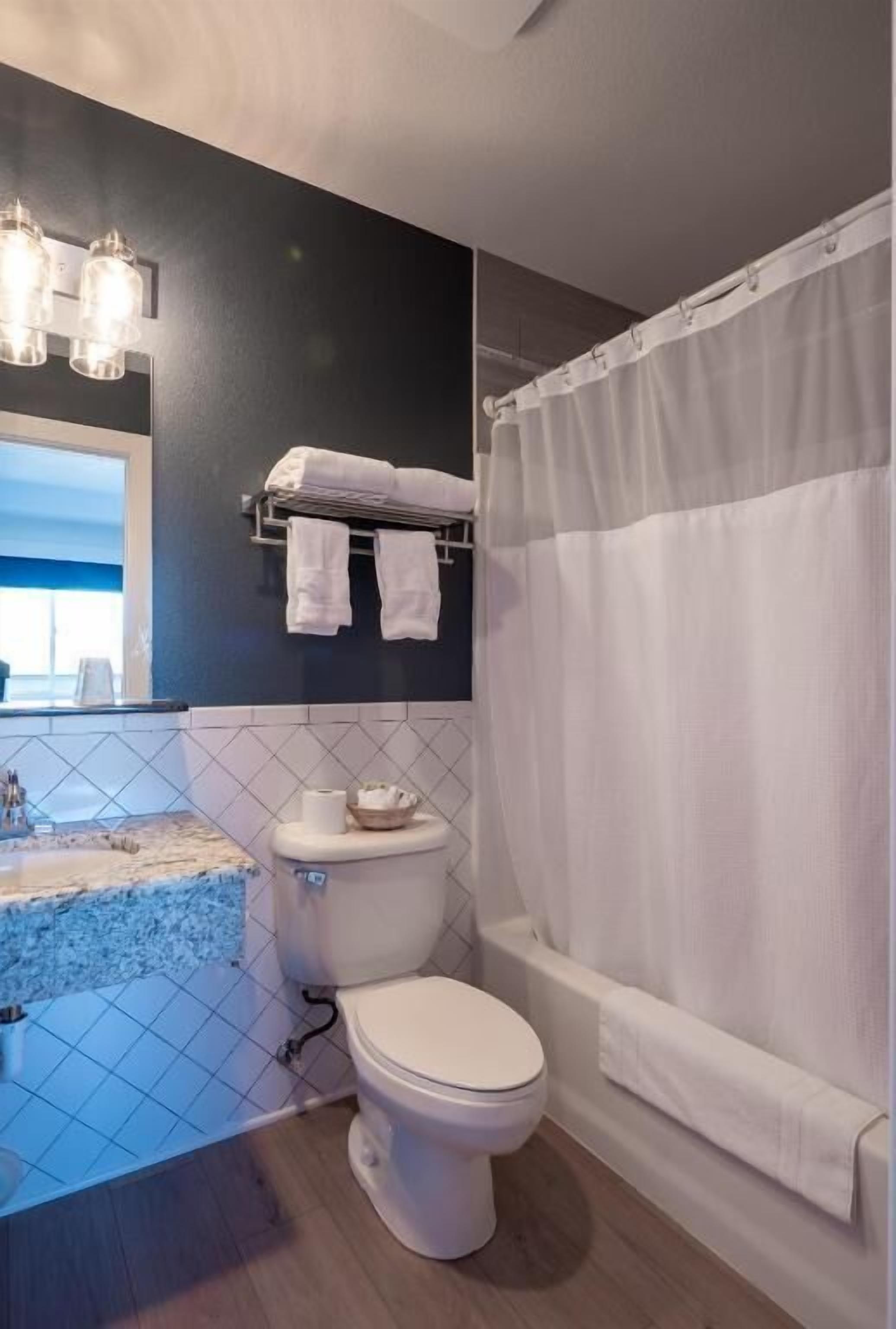 A bathroom with a toilet, a shower curtain, a towel rack with towels, and a sink with a tiled backsplash.