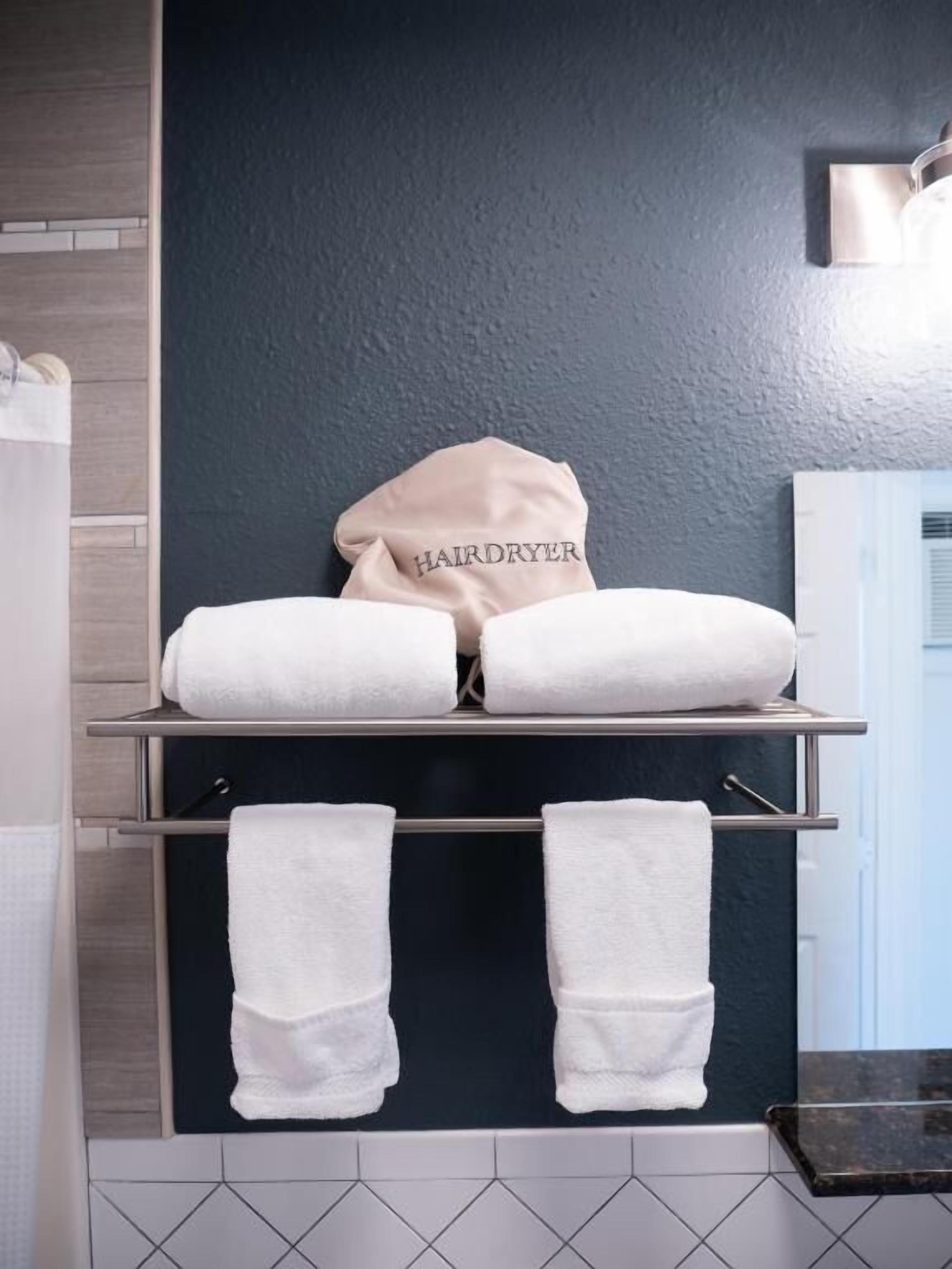 A bathroom with a towel rack holding two towels and a hairdryer bag labeled "HAIRDRYER" on a shelf.