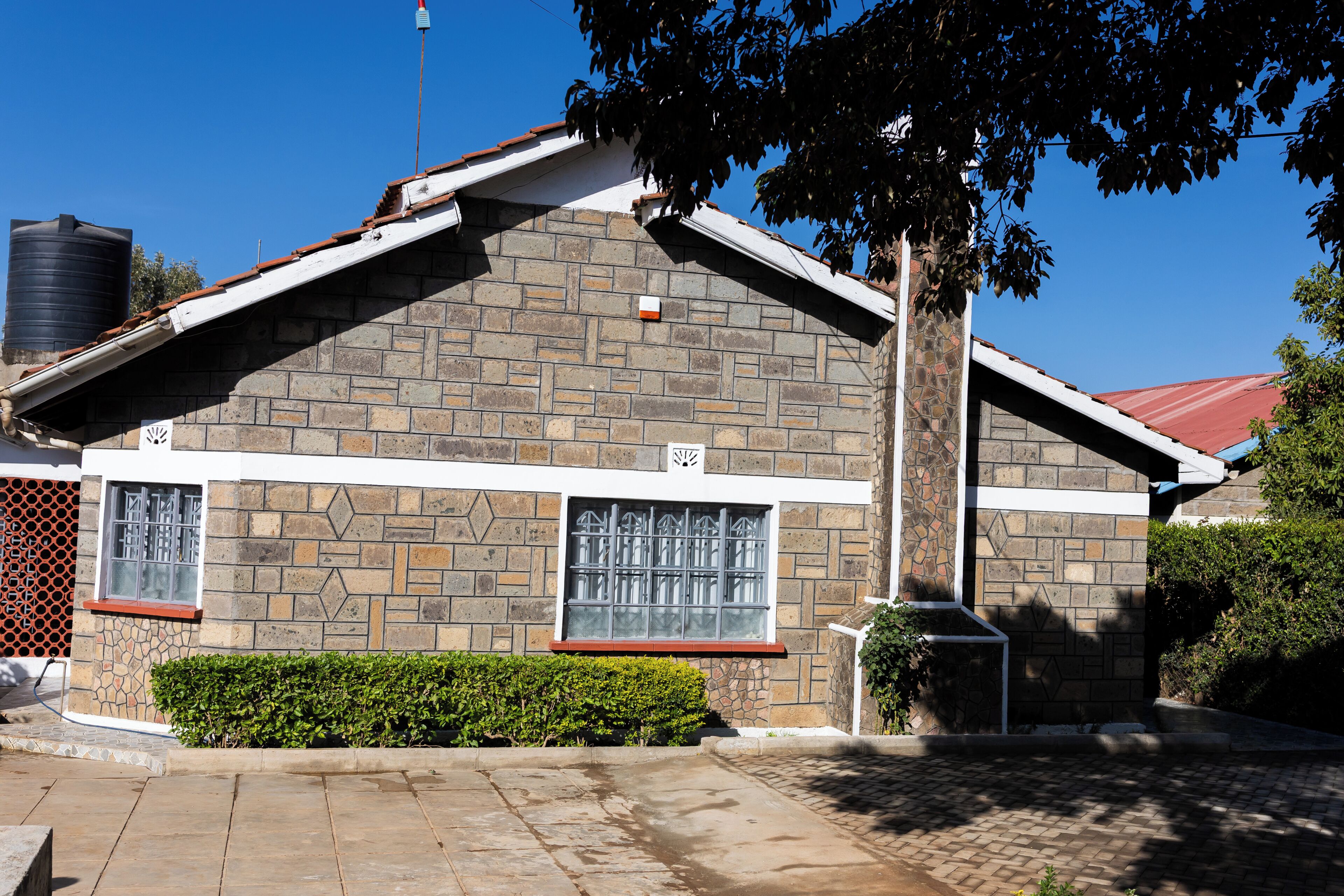 A two-story building with a stone facade, two windows with metal bars, a red roof, and a water tank in the background.