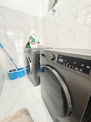 A laundry area with a front-loading washing machine, a blue mop bucket, and a bottle of Ariel Power Gel.