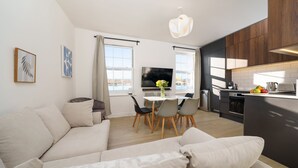 A modern living room with a sofa, dining table, and chairs, a kitchen with wooden cabinets, and a TV on the wall. - Luxury duplex with 3beds/3baths. 15mins to Arsenal FC and Camden Town & Market (London)