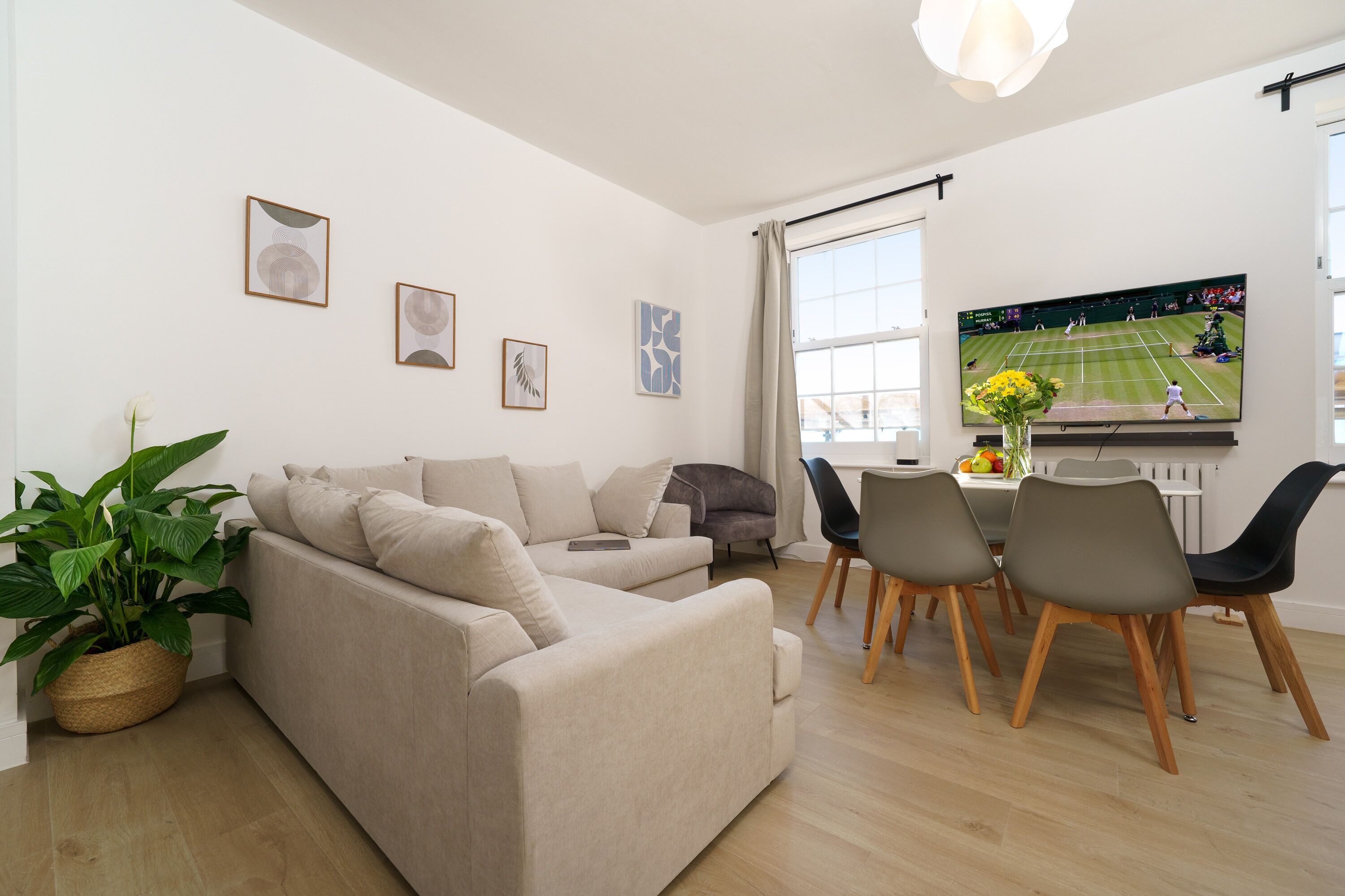 A modern living room with a beige sofa, dining table with chairs, and a flat-screen TV displaying a tennis match.