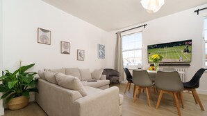 A modern living room with a beige sofa, dining table with chairs, and a flat-screen TV displaying a tennis match. - Luxury duplex with 3beds/3baths. 15mins to Arsenal FC and Camden Town & Market (London)