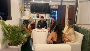 A group of people seated on a white couch in a room with a television mounted on the wall, a bar area with bottles, and a potted plant. - Hostal Mirador (Pisco)