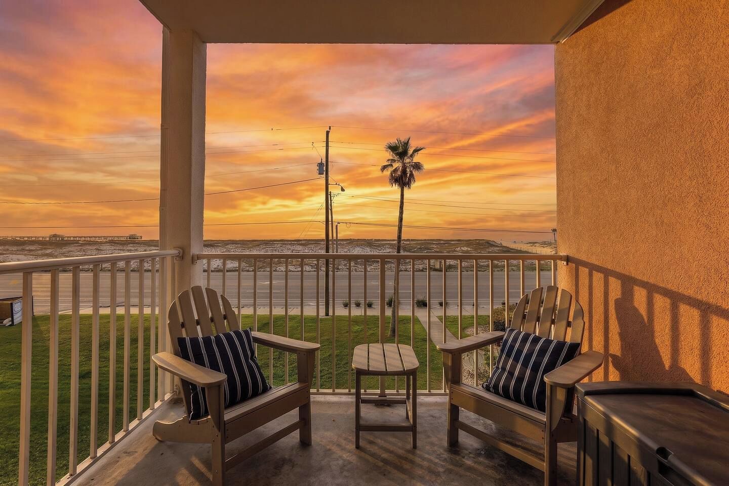 Een balkon met twee Adirondack-stoelen en een tafeltje, met uitzicht op een strand bij zonsondergang.