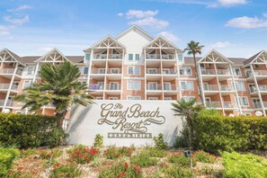 A multi-story hotel with balconies, a sign reading "The Grand Beach Resort Condominium," and landscaped gardens with red flowers. - Hot Tub & Pool Gulf Views King Bed Gulf State Park (Gulf Shores)
