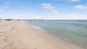 Ein Strand mit einem Steg, der sich ins Wasser erstreckt, unter blauem Himmel, während Menschen das Wasser genießen.