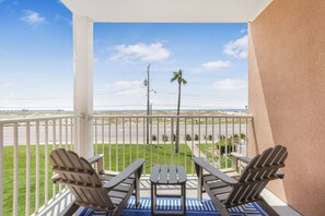 A balcony with two Adirondack chairs and a small table, overlooking a grassy area with a road and palm trees. - Family Beach Getaway Gulf View Balcony Large Pool, Hot Tub & King Bed (Gulf Shores)
