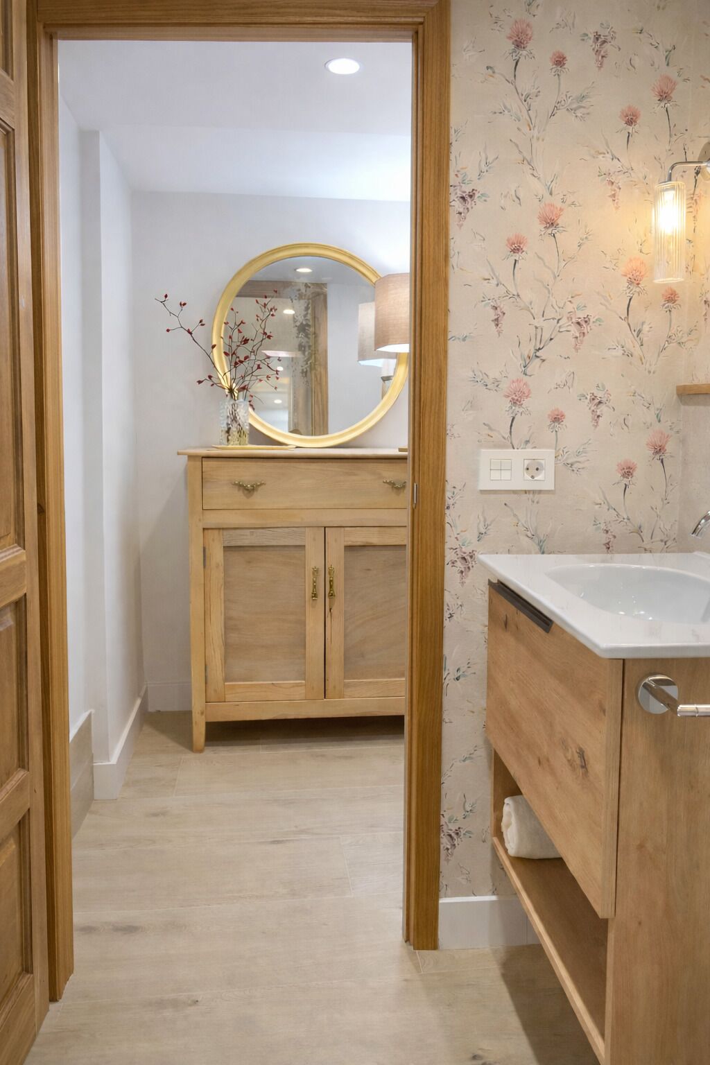 Een badkamer met een houten wastafelmeubel, een ronde spiegel en bloemenbehang.