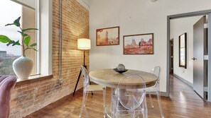 A dining area with a brick wall, a round table, clear chairs, a vase, and two paintings. - Downtown Dallas - Sleeps 6 People! - Gym - Pool (Dallas)