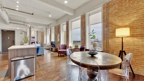 Uma sala de estar ampla com uma mesa de jantar de madeira, cadeiras transparentes e uma grande janela com vista para a cidade.