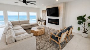 A modern living room with a large sofa, a coffee table, a fireplace, a TV, and a view of the ocean.