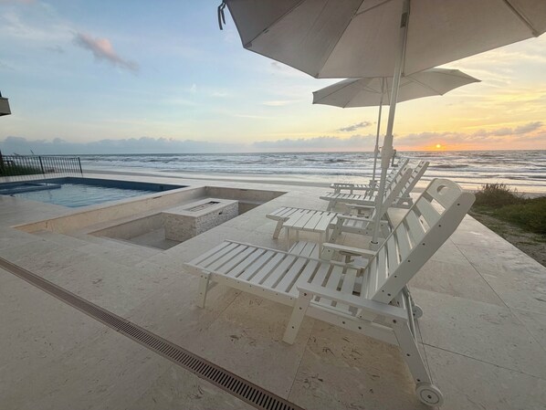 Zona de piscina con sillones de descanso y una sombrilla, con vista al océano al atardecer.