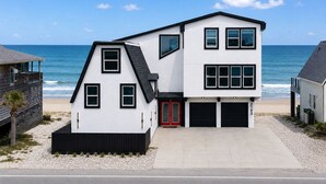 A modern two-story house with a garage, located on a beach with the ocean in the background.
