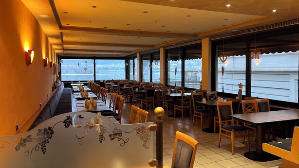 Un restaurante con una barra larga, varias mesas y sillas. Amplias ventanas ofrecen vistas al exterior.