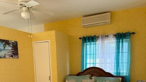 Une chambre à coucher comprenant un lit, un ventilateur de plafond, un climatiseur, une table de chevet et un mur orné d’un tableau.