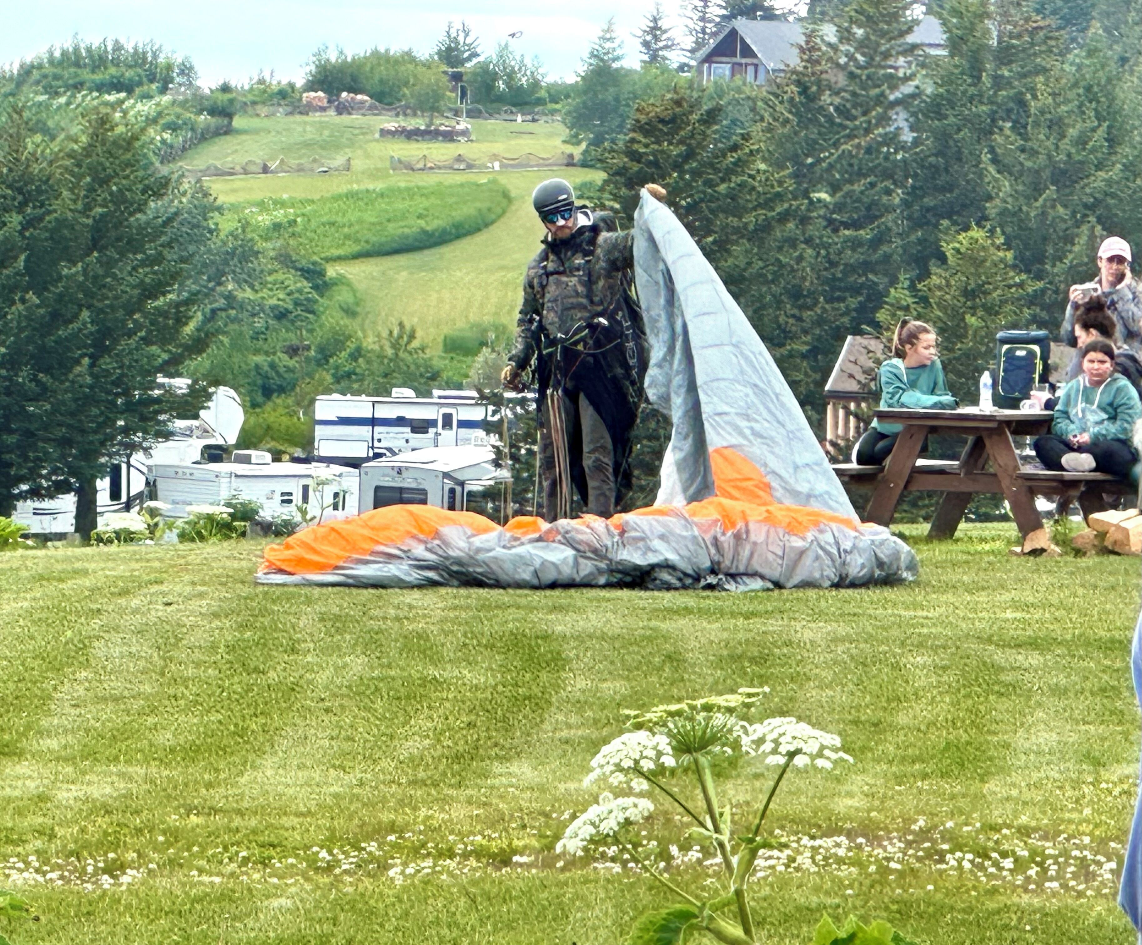Ein grasbewachsenes Feld mit einer großen, fallschirmartigen Konstruktion, einem Picknicktisch und einigen Personen im Hintergrund.