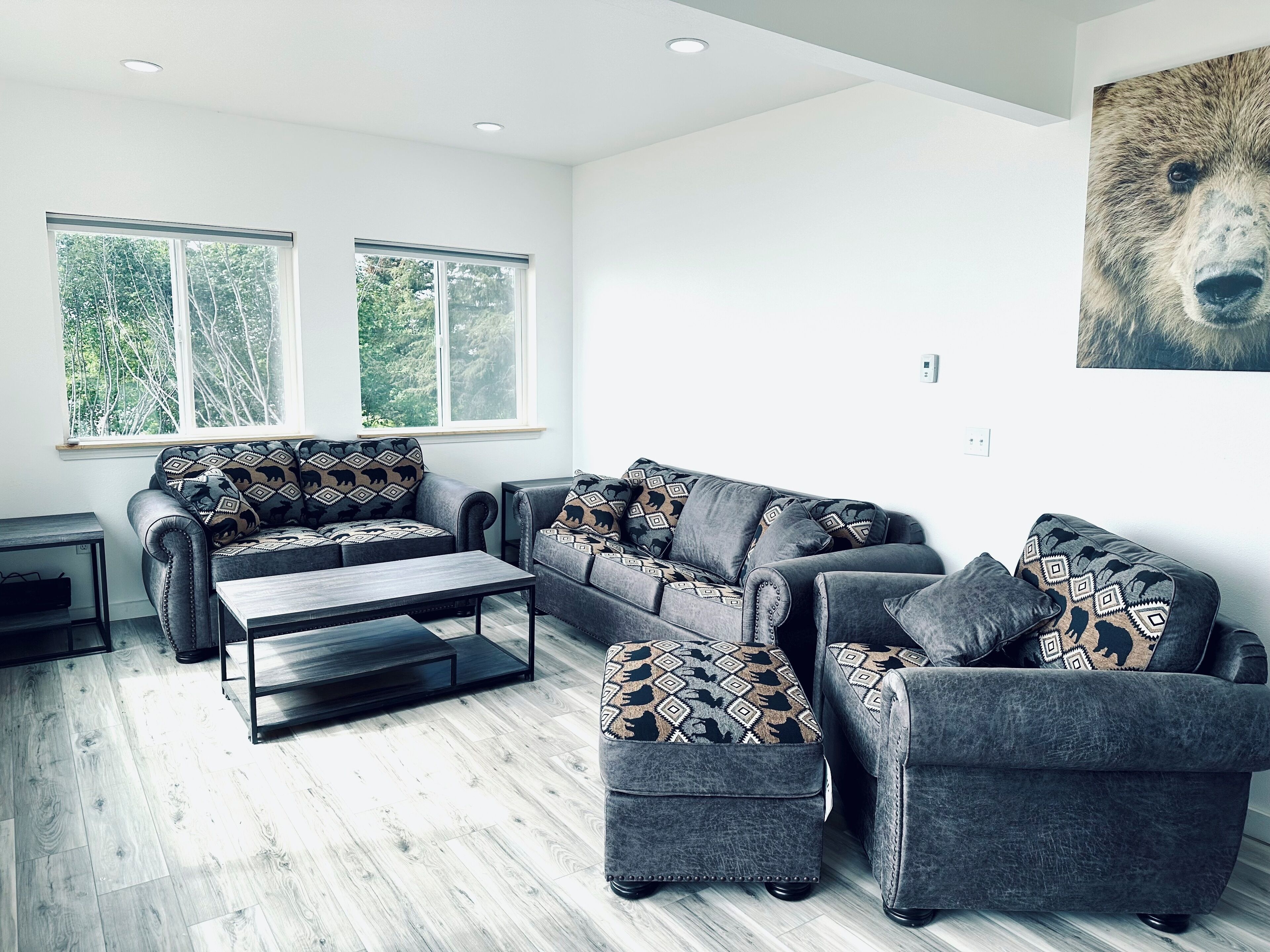 A modern living room with a grey sectional sofa, matching ottoman, and a coffee table. There are two windows providing natural light and a view of greenery outside.