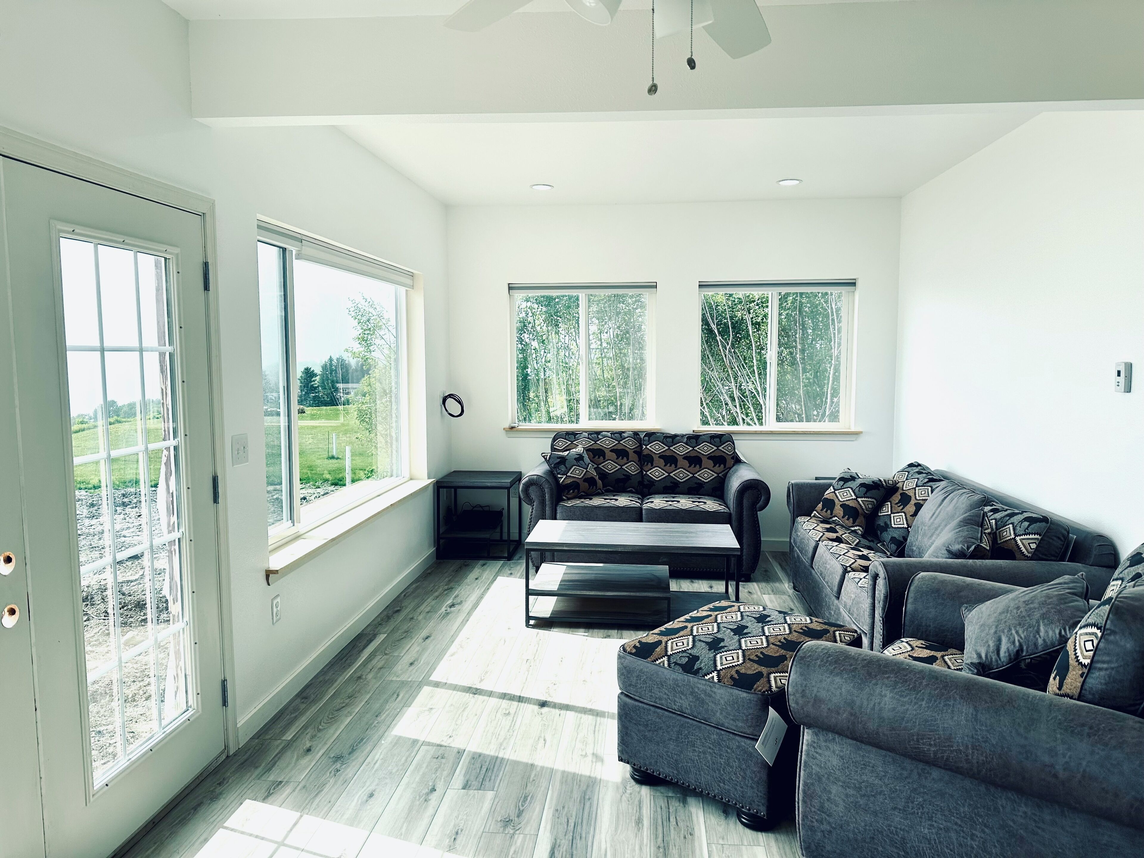 A spacious living room with a sofa, ottoman, and a coffee table. There are large windows providing natural light and views of greenery outside.