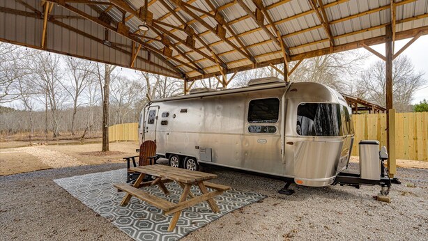 Une vieille caravane est garée sous un abri, à proximité d’une table de pique-nique et de toilettes portables.