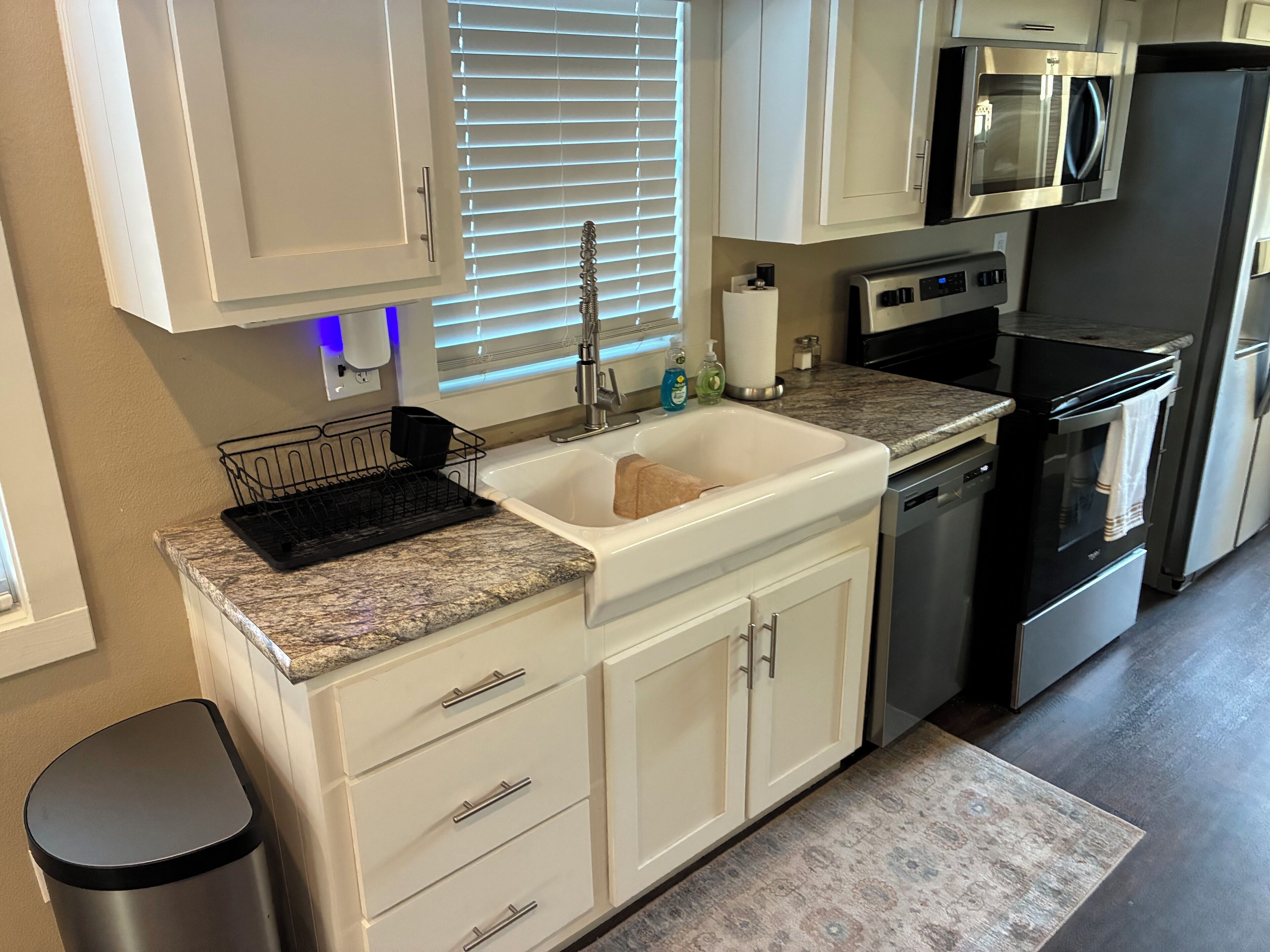 A modern kitchen with white cabinets, a granite countertop, a stainless steel sink, a microwave, an oven, and a trash can.