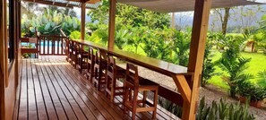 Una terraza de madera con una larga mesa y sillas, rodeada de abundante vegetación y vista a una piscina.
