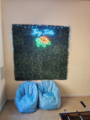 A room with a wall-mounted neon sign reading 'Tipsy Turtle' and a turtle graphic, a blue bean bag chair, and a wooden table with a remote control.