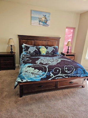 A bedroom with a large bed, a nightstand, a lamp, a window with blinds, and a painting on the wall.