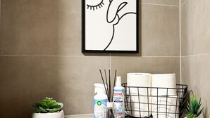 Casa de banho com um desenho de um pássaro emoldurado, uma pequena planta em vaso, um frasco de spray de higiene e uma cesta de toalhas.