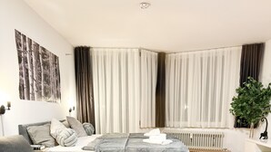 Quarto moderno e bem iluminado, com um sofá cinza, uma cama com cobertura cinza e uma mesa branca.