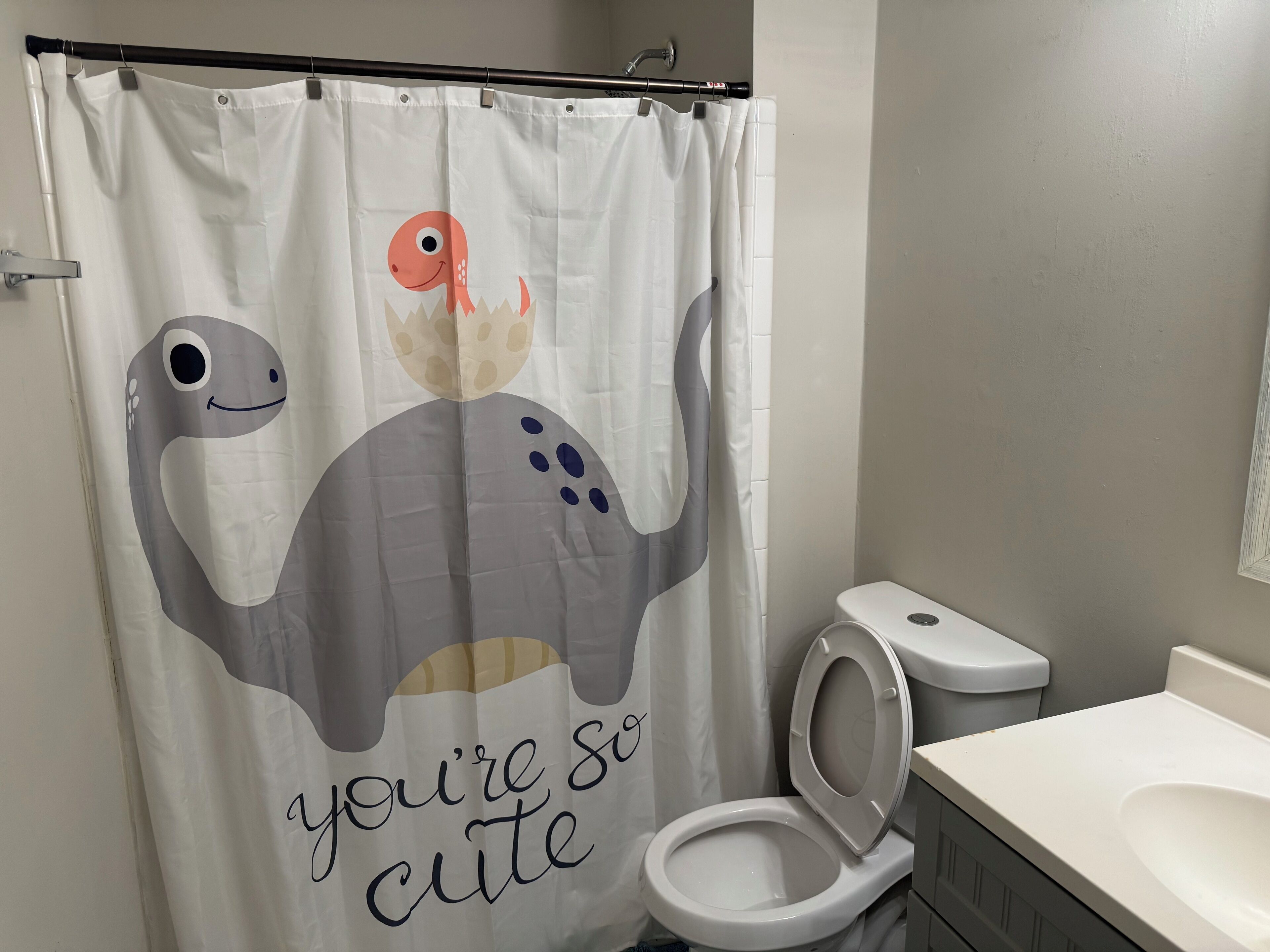 Ein Badezimmer mit Toilette, Waschbecken und einer Duschvorhang mit Dinosaurier-Motiv und dem Schriftzug 'you're so cute'.