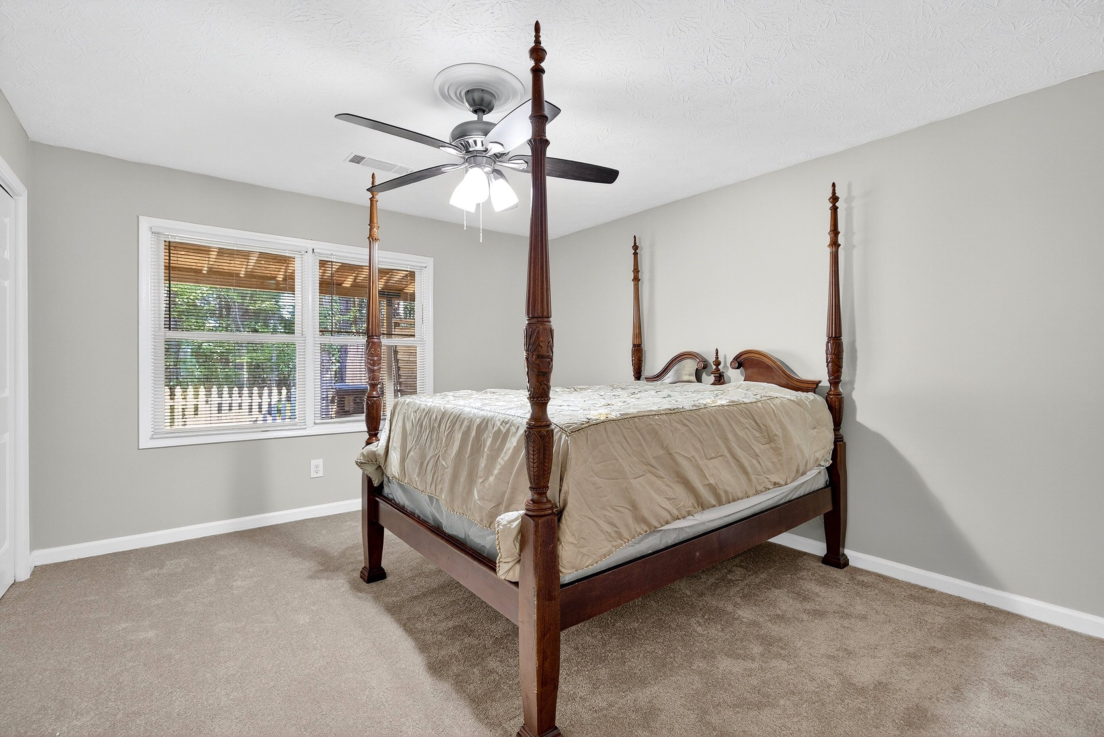 A four-poster bed with a ceiling fan, a window with blinds, and a carpeted floor.
