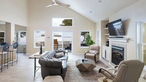 Un salón amplio con techo alto, chimenea, televisor montado y un gran ventanal con vistas al exterior.