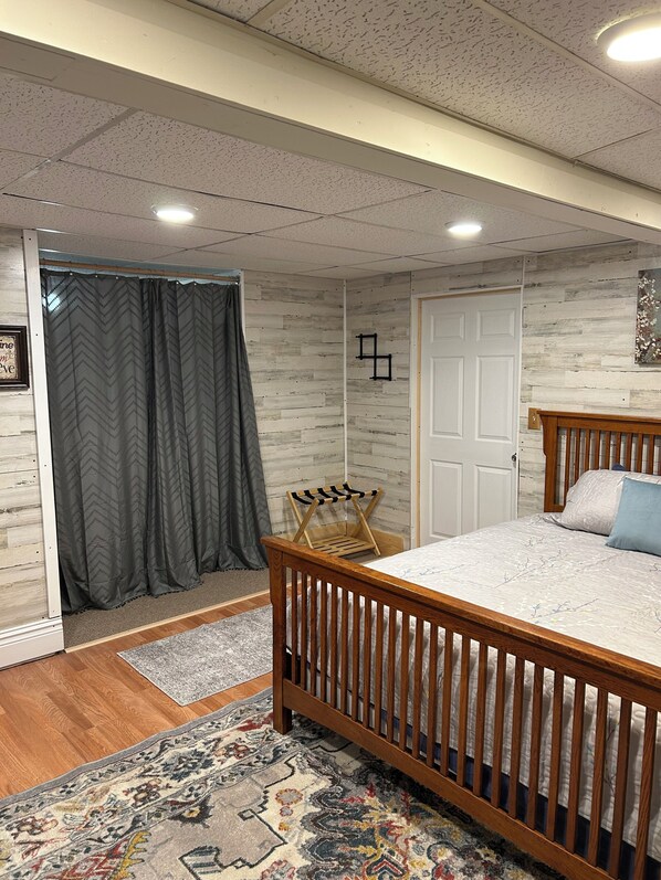 A bedroom with a wooden bed, a gray patterned curtain, a small wooden chair, and a patterned rug.