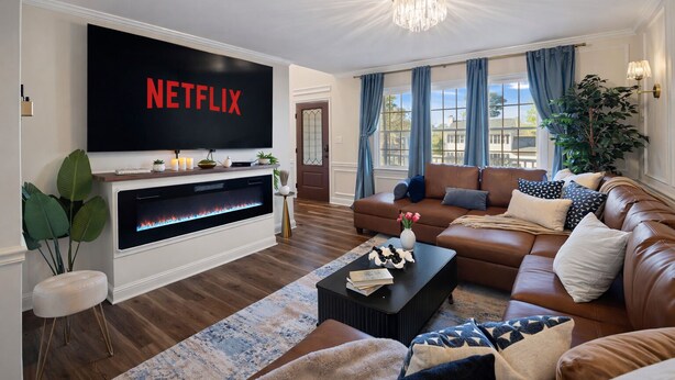 A modern living room with a fireplace, a large TV displaying 'NETFLIX', a leather sofa, and a chandelier.