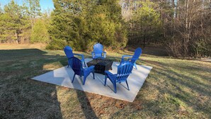 A concrete patio with four blue Adirondack chairs and a square table, surrounded by a grassy area with trees in the background.