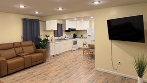 A modern living room with a brown leather sofa, a dining area with a table and chairs, a kitchen with white cabinets and appliances, and a mounted flat-screen TV.