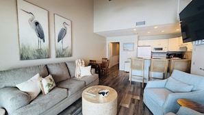 A spacious living area with a grey sofa, a round woven ottoman, and a light blue sofa. There are two framed bird paintings on the wall, a kitchen area with white cabinets, and a ceiling fan.