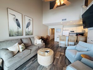 Un vaste espace de vie comprenant un canapé gris, un pouf rond en fibres tressées et un canapé bleu clair. Deux tableaux représentant des oiseaux, encadrés, ornent le mur. On y trouve également un coin cuisine avec des meubles blancs et un ventilateur de plafond.