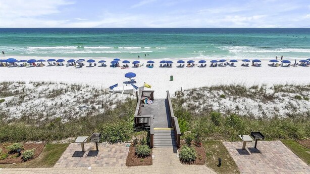 A beach with rows of blue umbrellas and lounge chairs, a wooden walkway leading to the beach, and a grassy area with a small structure.