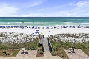 A beach with rows of blue umbrellas and lounge chairs, a wooden walkway leading to the beach, and a grassy area with a small structure.