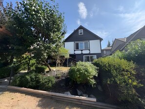 Ein zweistöckiges Haus mit dunkler Fassade, einem sichtbaren Fenster und einem Garten mit verschiedenen Pflanzen und einem kleinen Baum.