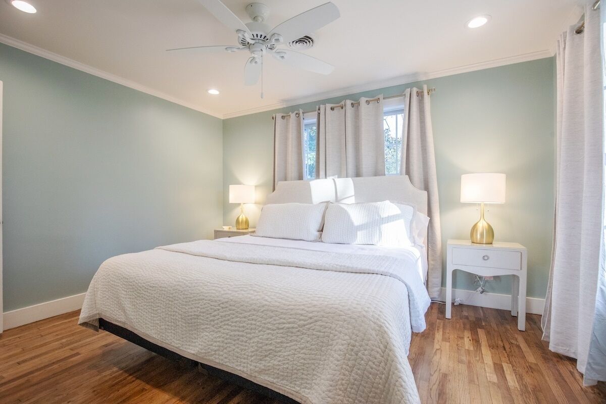 A bedroom with a large bed, two bedside tables with lamps, a ceiling fan, and a window with curtains.